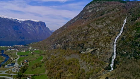 Toma-En-ángulo-Alto-De-La-Hermosa-Cascada-Por-Las-Montañas-Rocosas-En-Aurland,-Noruega-Con-La-Vista-De-Las-Casas-De-La-Ciudad-Debajo-Durante-El-Día