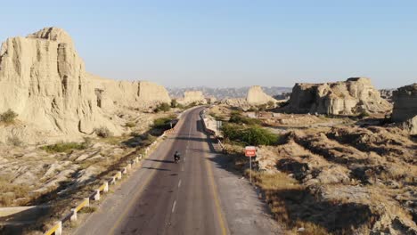 Tiro-De-Seguimiento-Aéreo-Detrás-De-La-Conducción-De-Motocicletas-A-Lo-Largo-De-Una-Carretera-Vacía-A-Través-Del-Parque-Nacional-Hingol-En-El-Paisaje-Desértico-De-Baluchistán