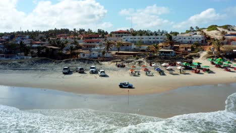 Aerial-dolly-out-drone-shot-of-a-beautiful-tropical-beach-in-Sibauma-near-Pipa-in-Northern-Brazil-with-tourist-umbrellas,-golden-sand,-clear-water,-sand-buggies,-and-beach-homes