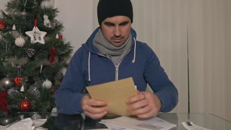 Cold-desperate-male-confused-and-upset-sorting-through-pile-of-bills-at-Christmas