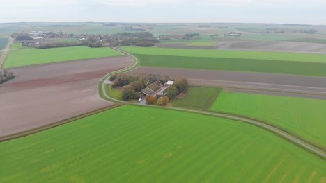 High-drone-shot-above-the-grass-green,-brown,-earthy-farmlands-and-farmhouse-of-the-Netherlands