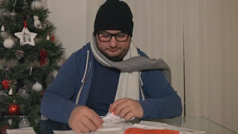 Cold-male-wearing-hat-and-glasses-frustrated,-upset-with-pile-of-finance-receipts-at-Christmas