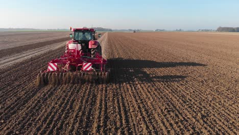 Red-tractor-plowing-brown,-earthy,-dutch-farmland-in-vertical-lines-under-blue-skies
