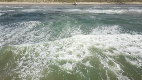 The-waves-from-the-Gulf-of-Mexico-break-onto-the-beach-in-Port-Aransas,-Texas-while-a-truck-full-of-people-enjoy-sandy-shores