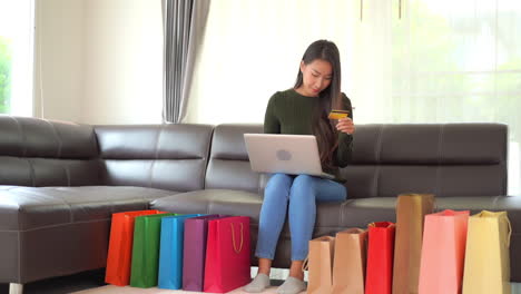 Glückliche-Asiatische-Frau,-Die-Online-Mit-Kreditkarte-Und-Laptop-computer-Mit-Vollen-Bunten-Taschen-Auf-Dem-Boden-Im-Wohnzimmer-Einkauft