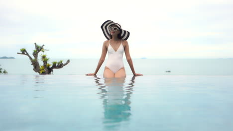 Schöne-Asiatische-Frau,-Die-In-Badeanzug,-Sonnenbrille-Und-Sommerhut-Am-Rand-Des-Infinity-Pools-Sitzt