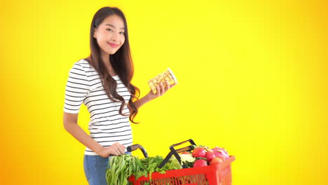 Slim-woman-reading-a-label-of-a-snack-while-shopping-for-food-and-vegetables-on-isolated-yellow-background