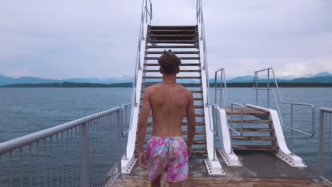 Slow-motion-shot-of-a-young-man-walking-towards-diving-board-and-walking-up-the-stairs,-watching-over-beautiful-seascape