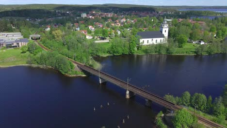 Train-bridge-over-small-blue-water-lake-with-a-white-church-embedded-in-green-trees,-in-the-background-the-part-of-the-city-is-visible