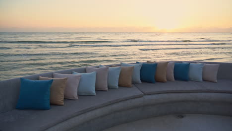 Ocean-waves-roll-toward-the-shore-in-the-background,-foreground-an-empty-padded-sitting-area-covered-end-to-end-with-colorful-pillows