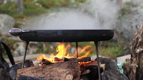 Close-up-shot-of-muurikka-traditional-finish-classic-griddle-pan,-over-a-hot-wood-fire-and-steaming-water-being-poured-over,-in-a-camping-environment