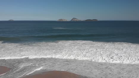 Sideways-aerial-view-of-waves-coming-in-on-Ipanema-beach-in-Rio-de-Janeiro-with-islands-just-outside-the-coast-in-the-background-on-a-sunny-afternoon-with-pristine-sand-beach-and-few-people-for-scale