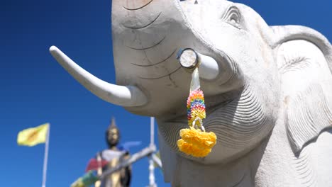 Statue-Von-Ganesha,-Elefant,-Mit-Blumenopfer-Auf-Stoßzähnen,-Gebetsfahnen-Und-Buddha-Statue-Im-Hintergrund