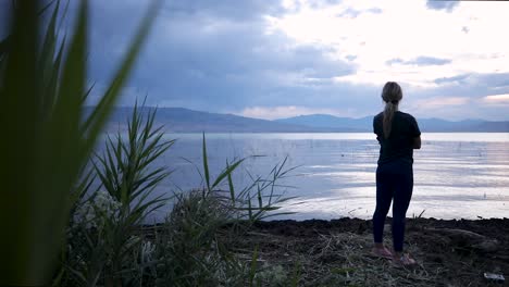 Young-Woman-standing-alone-at-calm-waterfront-lake-with-mountain-range-in-background-on-cloudy-day,-handheld-pan-slow-motion