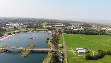 Drone-rising-above-lake-over-seaside-town,-people-walking-across-the-bridge-on-sunny-day