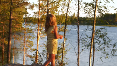 Blonde-girl-standing-between-trees-looking-out-at-lake,-behind