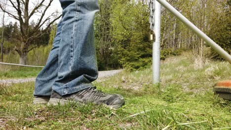 Ankle-view-of-person-wearing-jeans-cutting-green-grass-with-weed-eater-by-metal-steel-poll-in-countryside