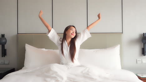 Attractive-asian-female-waking-up-in-her-bed-with-smile-and-stretching-her-hands,-static-wide-view