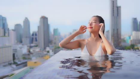 An-urban-skyline-frames-an-attractive-young-woman-at-the-edge-of-an-infinity-pool-sweeps-her-wet-hair-back-from-her-face,-Bangkok,-Thailand