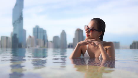 Attractive-wealthy-asian-female-with-sunglasses-in-infinity-rooftop-pool-of-luxury-hotel-with-blurred-cityscape-in-background,-full-frame-slow-motion