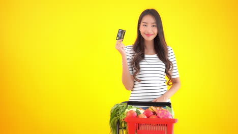 A-young-healthy-woman-with-a-basket-of-healthy-food-holds-up-a-credit-card-and-point-to-it