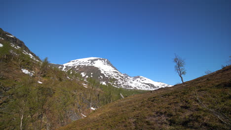 Antenne,-Schwenkdrohnenaufnahme-Von-Kahlen-Hügeln-Und-Schneebedeckten-Berggipfeln-Im-Hintergrund,-Blauer-Himmel,-An-Einem-Sonnigen-Sommertag,-In-Den-Lyngenalpen,-Nordnorwegen