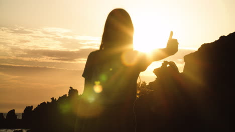 Young-girl-with-long-hair-silhouetted-by-rocky-landscape-gives-thumbs-up-with-bright-white-sun-and-glare-shining-in-background-sky,-static