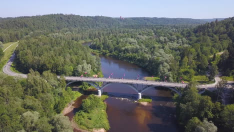 Hochwinkel-Luftaufnahme-Einer-Gruppe-Von-Radfahrern,-Die-Eine-Brücke-über-Einen-Fluss-In-Lettland-überqueren