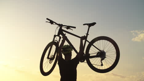 Caucasian-young-strong-man-lifts-mountain-bicycle-up-above-his-head-to-blue-sky-on-breezy-day,-static