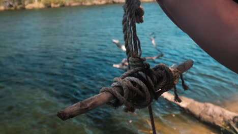 Close-Up:-hand-holds-the-end-of-the-rope-swing-with-wooden-sticks-on-the-river-bank
