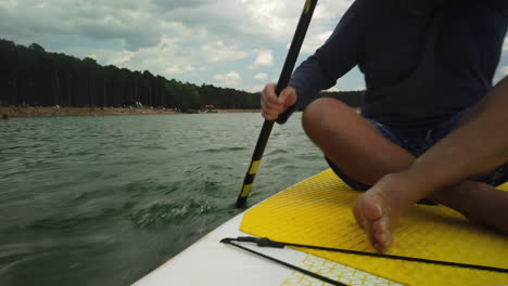 Man-paddles-fast-while-seated-on-paddleboard-past-lake-beach,-low-angle