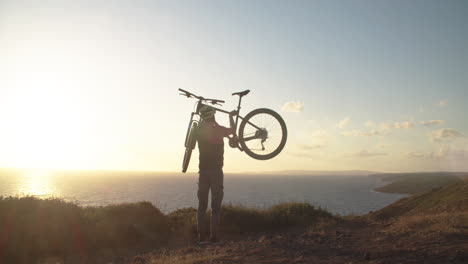 Young-Caucasian-cyclist-man-lifts-mountain-bicycle-up-above-his-head-in-countryside-by-blue-ocean-sea-with-bright-white-sun-in-background,-Sant'Antioco,-Sardinia,-Italy,-static