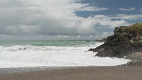 Zerklüftete-Küste-Und-Brechende-Wellen-Am-Strand-Mit-Wolken,-Die-In-Zeitlupe-An-Einem-Blauen-Himmel-Vorbeiziehen