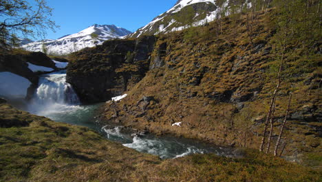 Statischer-Blick-Auf-Einen-Wasserfall-Mit-Schneebedeckten-Bergen-Im-Hintergrund,-An-Einem-Sonnigen-Sommertag,-In-Den-Lyngenalpen,-Nordnorwegen