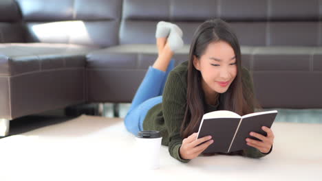 Happy-Asian-Female-Laying-on-Floor-of-Living-Room-and-Reading-The-Book,-Full-Frame-Slow-Motion