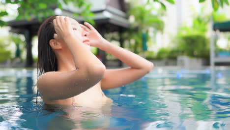 While-running-her-finger-through-her-long-wet-hair,-a-beautiful-young-woman-enjoys-the-waters-of-her-luxury-hotel-pool