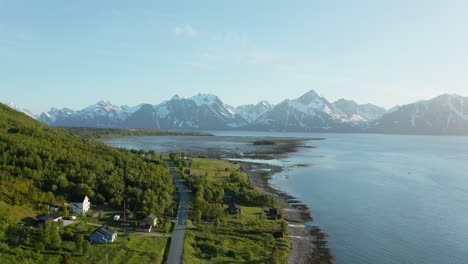 Aerial-view-of-houses-and-a-coastal-road,-the-Arctic-sea-and-snowy-mountain-fjord-in-the-background,-sunny,-summer-day,-in-Rotsund,-Troms,-Nordland,-Norway---rising,-drone-shot
