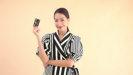 Asian-model-in-black-and-white-dress-posing-with-credit-or-debit-card-against-plain-background