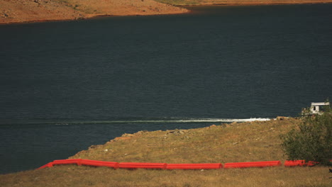 Boat-traveling-on-lake-near-Oroville-Dam-with-red-buoys-on-grass,-hot-summer-day