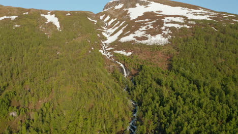 Aerial-view-around-a-waterfall-on-a-snowy-mountain,-sunny,-summer-day,-in-Rotsund,-Troms,-Nordland,-Norway---orbit,-drone-shot