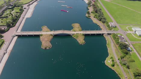 Drone-flying-over-bridge-on-lake-in-seaside-town,-people-walking-across-the-bridge-on-sunny-day