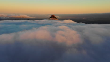 Aerial,-drone-shot-rising-over-clouds-towards-a-sunlit-mountain-peak,-during-sunset,-in-Hassell-National-Park,-West-Australia