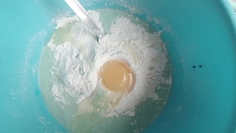 Homemade-baking-dropping-egg-into-sticky-raw-cake-batter-mixture-in-blue-bowl