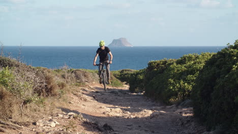 Man-rides-mountain-bike-up-dirt-narrow-rocky-path-in-countryside-by-the-coast,-Sardinia,-Italy,-static