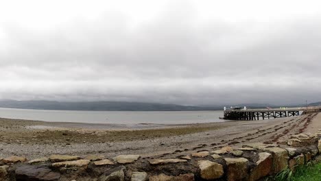 Historic-Beaumaris-pier-and-pebble-beach-timelapse-fast-clouds-above-misty-coastal-mountains