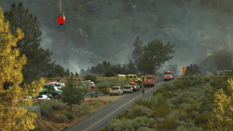 Helicopter-with-water-bucket-landing-on-road,-distant-view,-first-responders-on-site-for-wildfire,-firetrucks-with-flashing-lights,-Kern-River,-June-2020