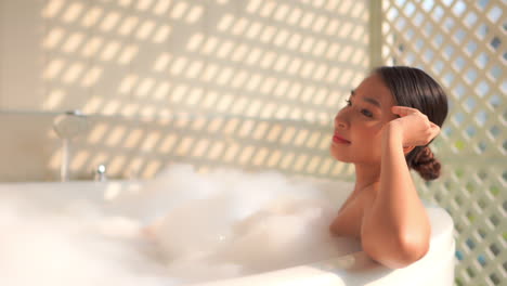 Attractive-young-Asian-woman-relaxing-in-outdoor-bath-filled-with-bubbles-as-soft-sunlight-shines-on-wall