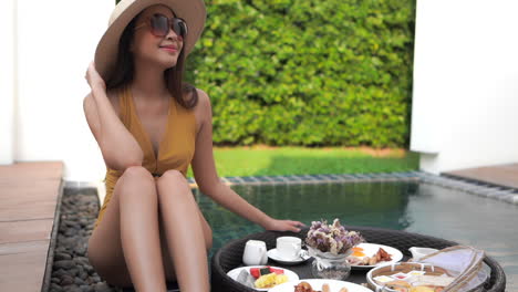 Woman-wearing-hat,-sunglasses-and-swimsuit-sitting-by-plunge-pool-with-breakfast-on-floating-tray