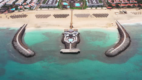 Aerial-shot-of-a-wide-sandy-beach-tourist-resort-with-a-twin-breakwater-construction,-sunbeds-and-umbrellas