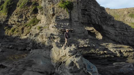 Aerial-orbit-around-a-bearded-man-standing-on-a-rocky-out-crop-underneath-coastal-cliffs